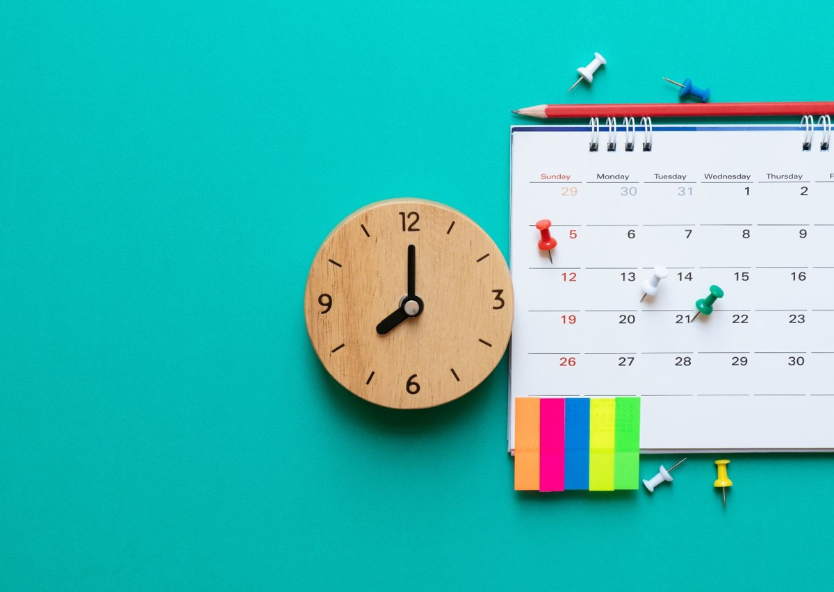 En kalender ligger mot en turkos bakgrund. Bredvid ligger en klocka i trä och post-it-lappar i glada färger.