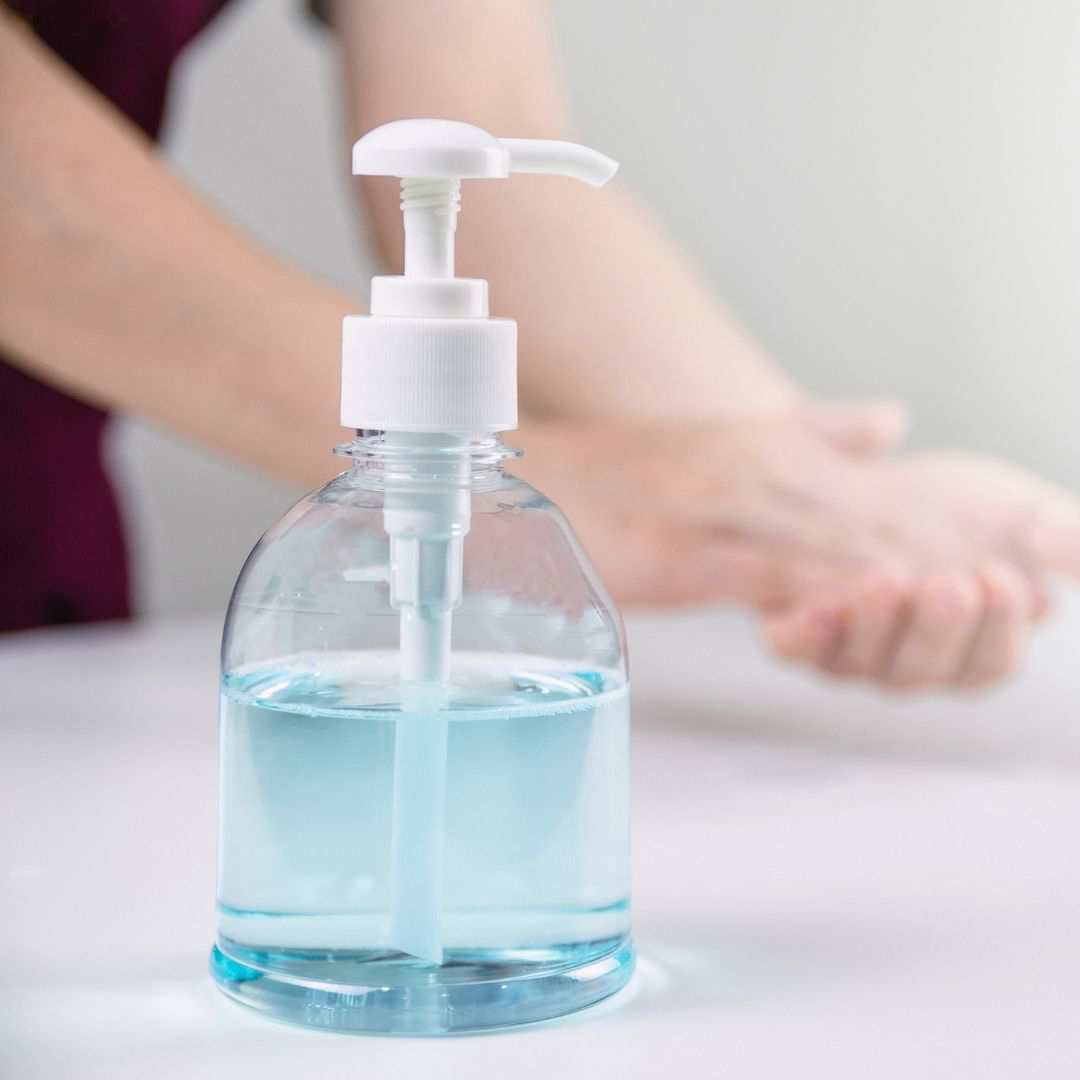 Foto på en genomskinlig pumpflaska med handsprit och i bakgrunden en manlig persons händer som tvättar sig.
