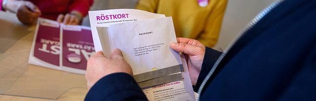 Foto på en person som håller upp sitt röstkort. I bakgrunden syns röstmottagarna lite ur fokus.