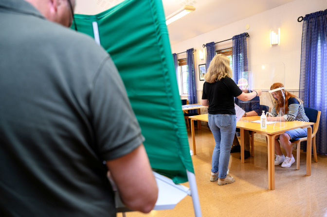 Foto på en person bakifrån som står bakom ett grönt valbås. I bakgrunden ser man en kvinna som står vid röstmottagarbordet.