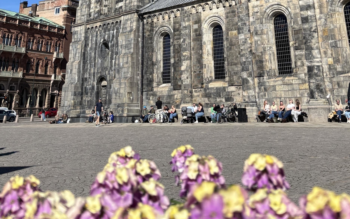 Människor sitter på bänkar längs med domkyrkan. Solen skiner.