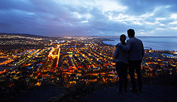 Två personer står på en höjd i mörkret och blickar ut över en upplyst stad.