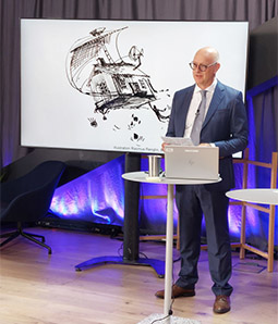 Konferensens moderator vid ett bord med dator och storbildsskärm i bakgrunden.