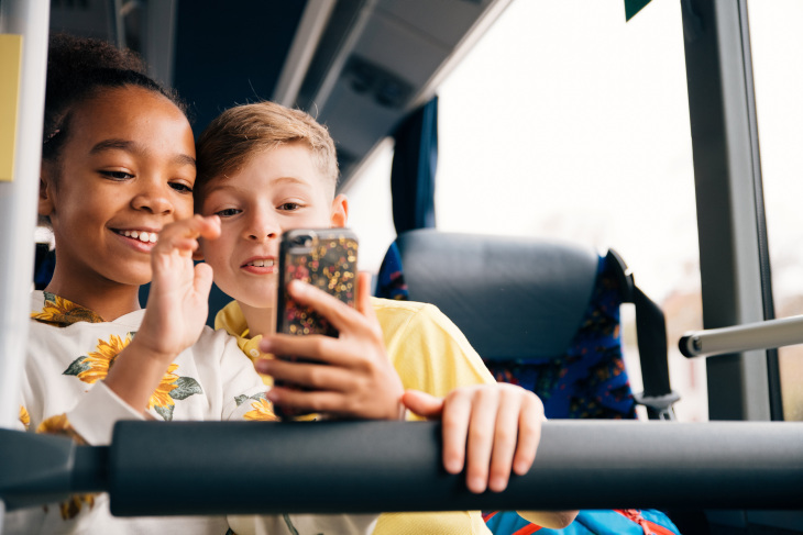 Två glada barn sitter i en buss och tittar på en smartphone