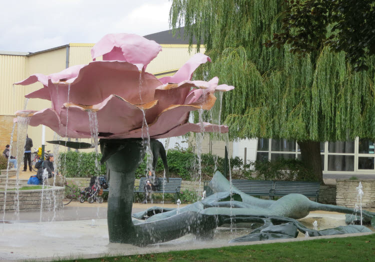 En skulptur som ser ut som en ros som sprutar vatten, i stadsmiljö med människor i bakgrunden.